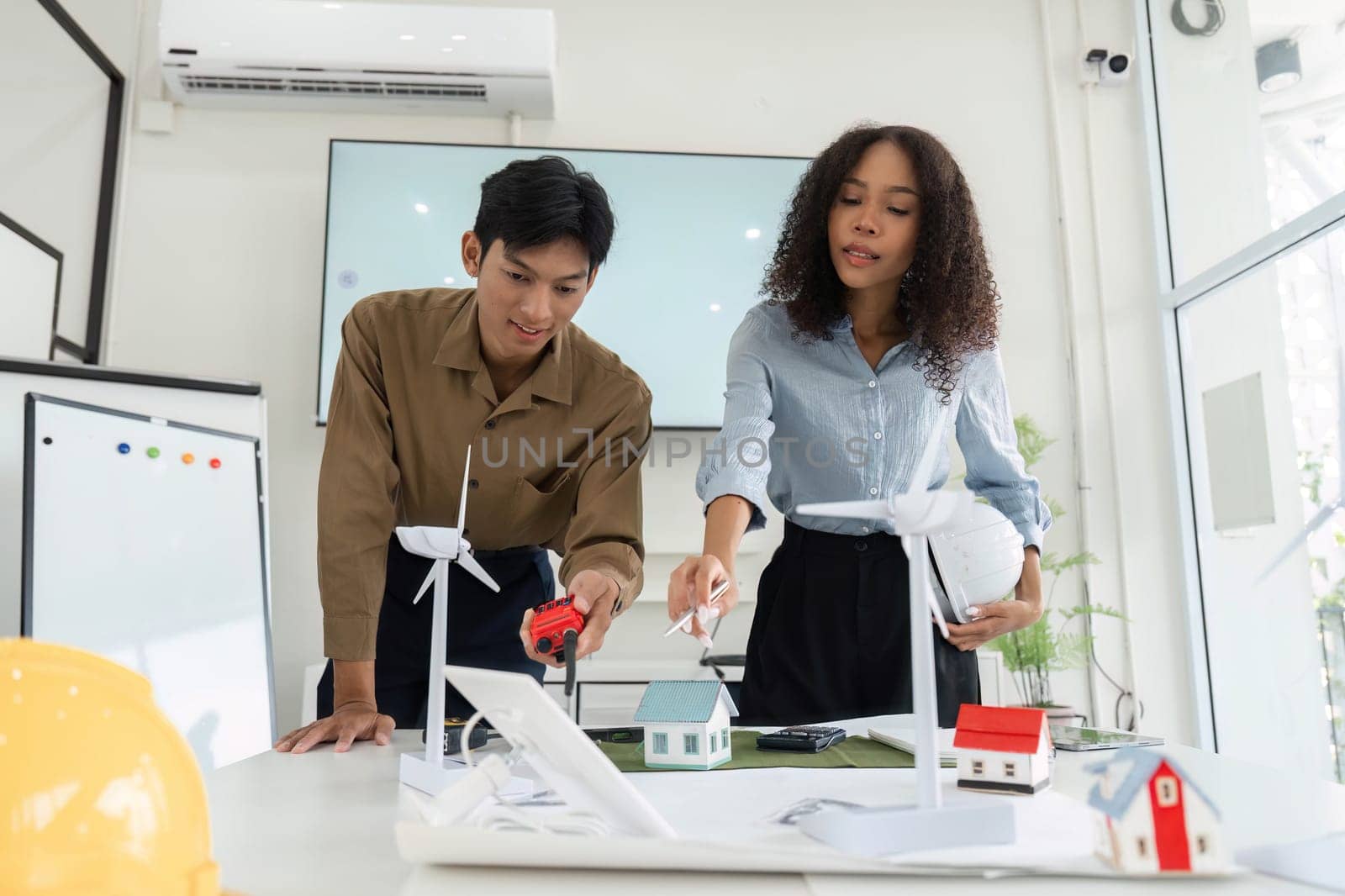 Two young professionals collaborate on sustainable housing solutions, showcasing wind turbines and model homes in a modern office environment.