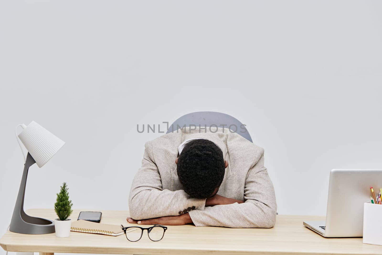 Tired man with dark hair and casual grey sweater, leaning forward with arms crossed on desk, displaying signs of mental exhaustion, in a modern workspace with minimal decor, capturing work stress and fatigue concept