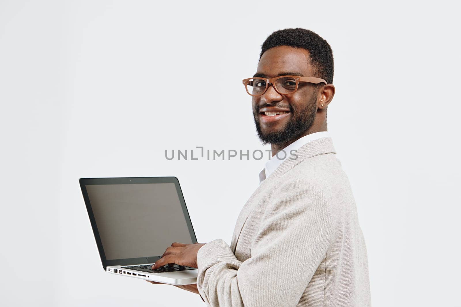 Happy man with glasses holding a laptop, dressed in a beige blazer and white shirt, smiling confidently, facing camera, studio setting with plain white background, representing technology, business, confidence, and professionalism, concept: modern work culture