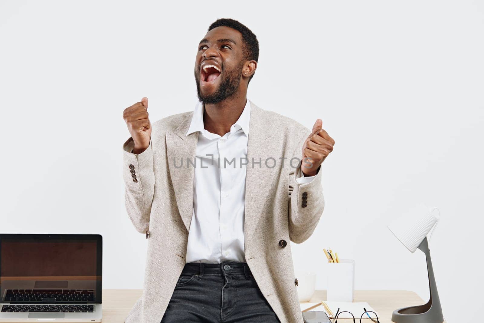 Happy man with a cheerful smile and raised fists, dressed in a beige blazer and white shirt, in a bright contemporary office environment, expressing excitement, success, motivation, modern work setting, positive energy. Concept: achievement and celebration.