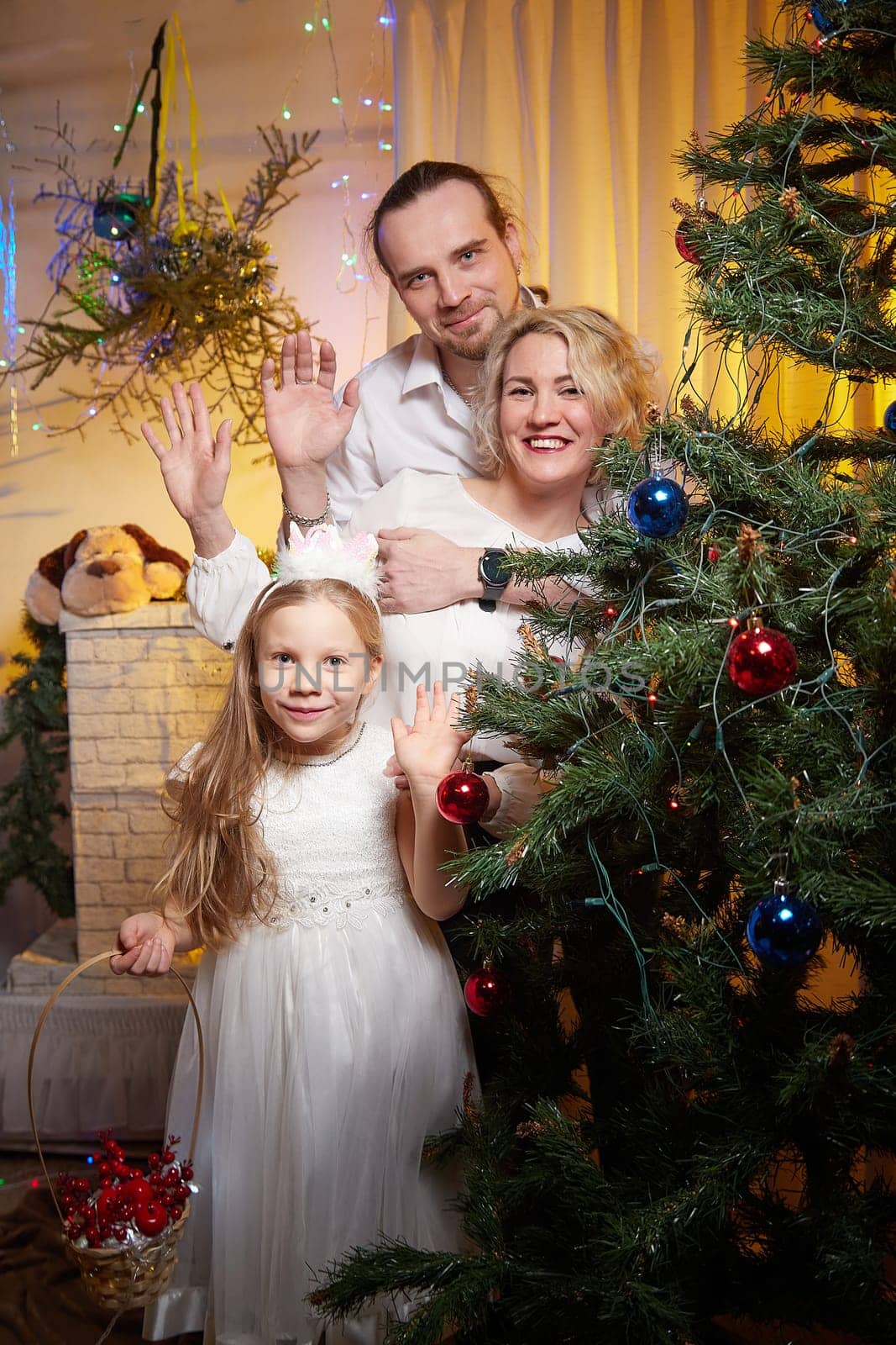 A family gathers in a warm room, smiling and waving near a festive Christmas tree adorned with colorful decorations and lights.