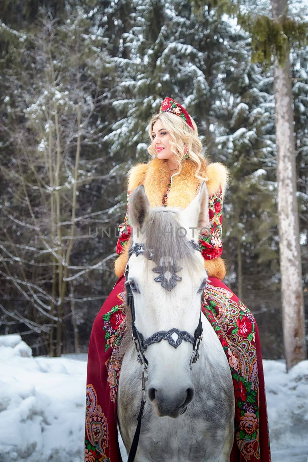A stunning Russian girl dressed in vibrant national attire enjoys a photoshoot with a horse amidst snowy scenery.