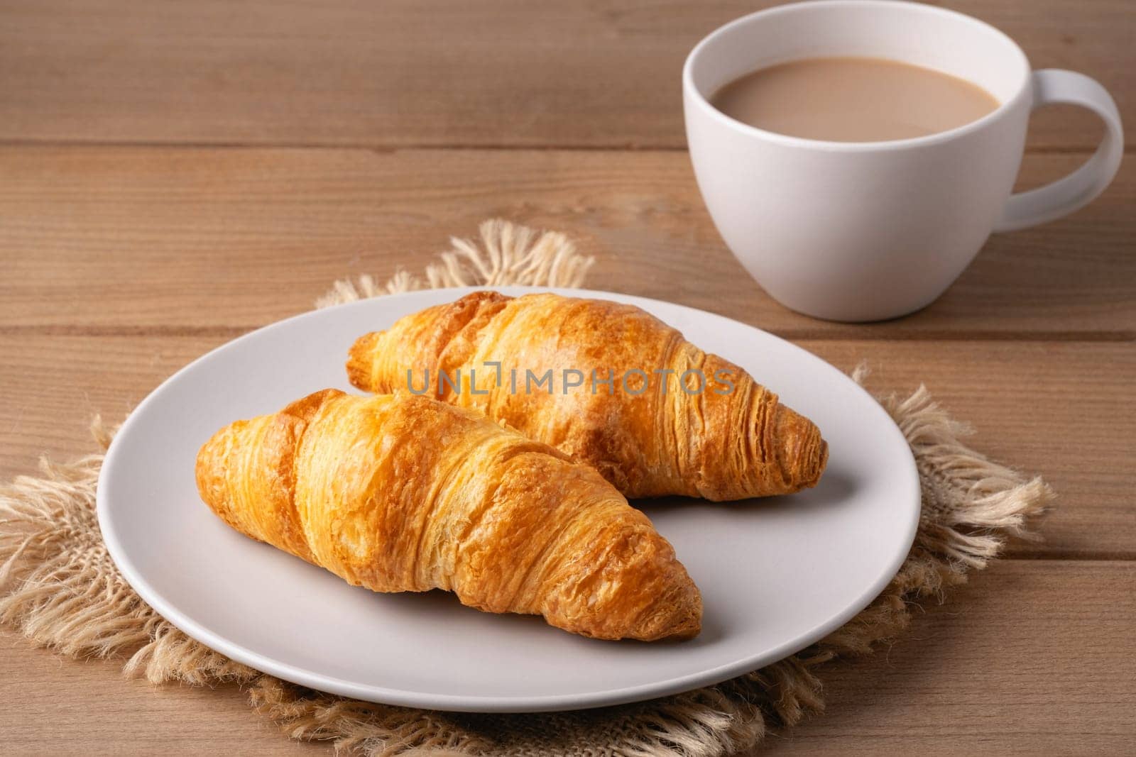 Two golden croissants rest on a white plate beside a steaming cup of coffee. The warm wooden table adds a rustic touch to this inviting breakfast arrangement.