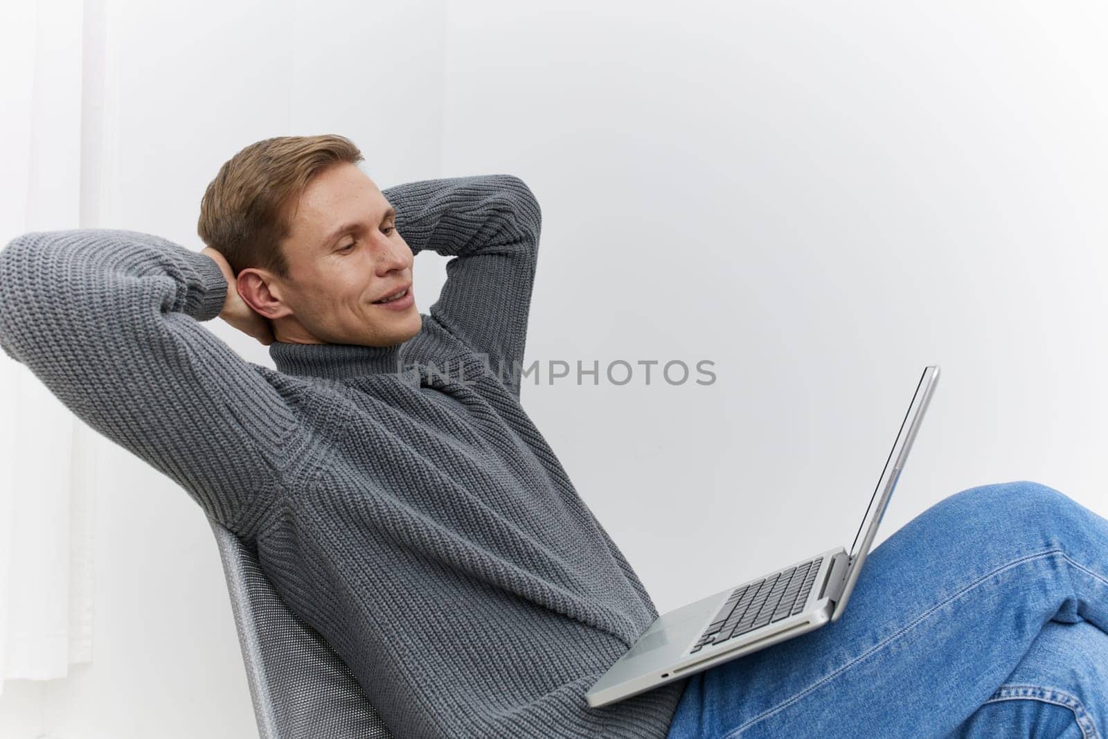 relaxed man using laptop while sitting in office chair wearing gray sweater and blue jeans focused on digital nomad lifestyle telecommuting remote work concept modern technology at home office space
