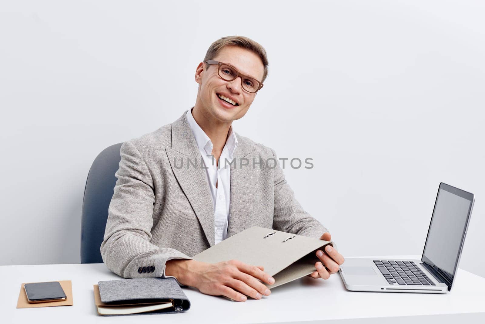 smiling man working with laptop at desk dressed in gray blazer and glasses using digital technology for freelance remote job as digital nomad concept with smartphone and notebook nearby