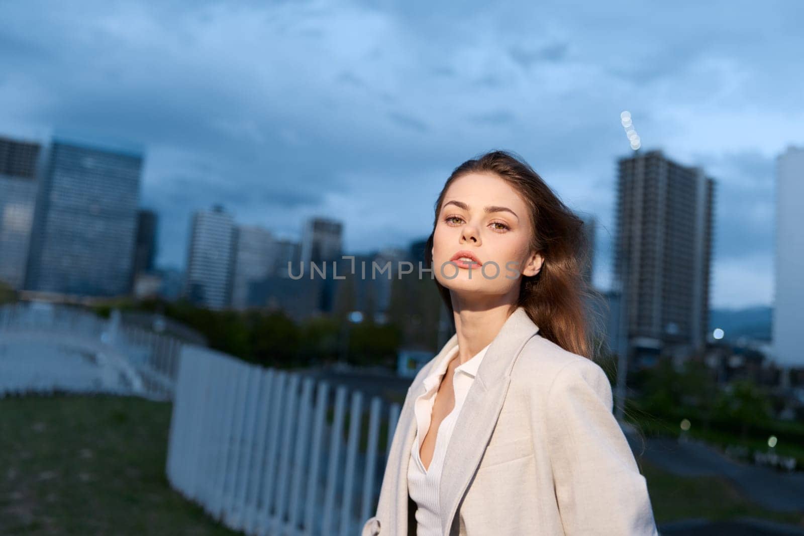 confident woman in beige jacket stands outdoors against a cityscape with modern buildings and cloudy sky at dusk, embodying urban lifestyle, empowerment, and professional business fashion.