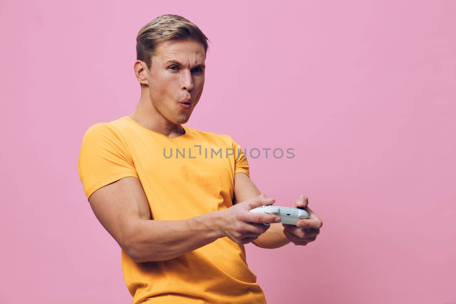 Cheerful man enjoying gaming session, wearing casual yellow t-shirt, holding game controller, showing surprise or excitement, isolated on pink background. Lifestyle entertainment shot