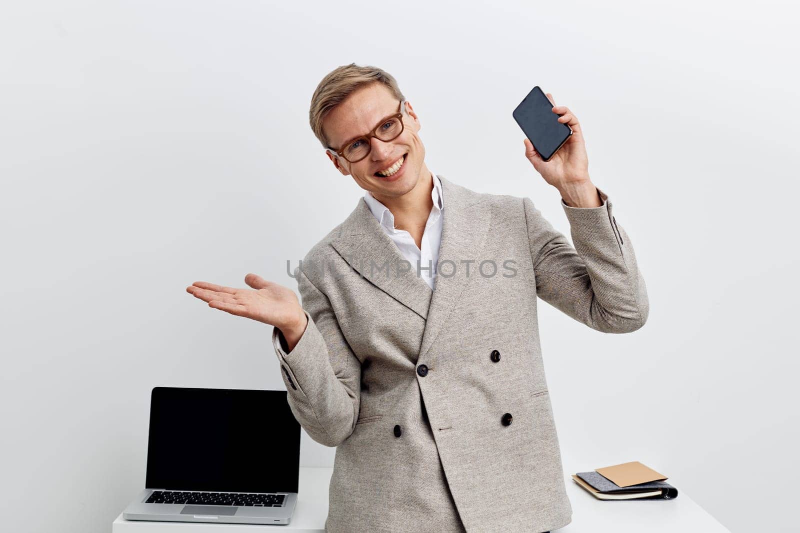 Happy young man wearing glasses and a light gray blazer poses with a smartphone, capturing a selfie in a bright studio setting. Casual yet professional style, positive mood, emphasizing confidence, modern business lifestyle.