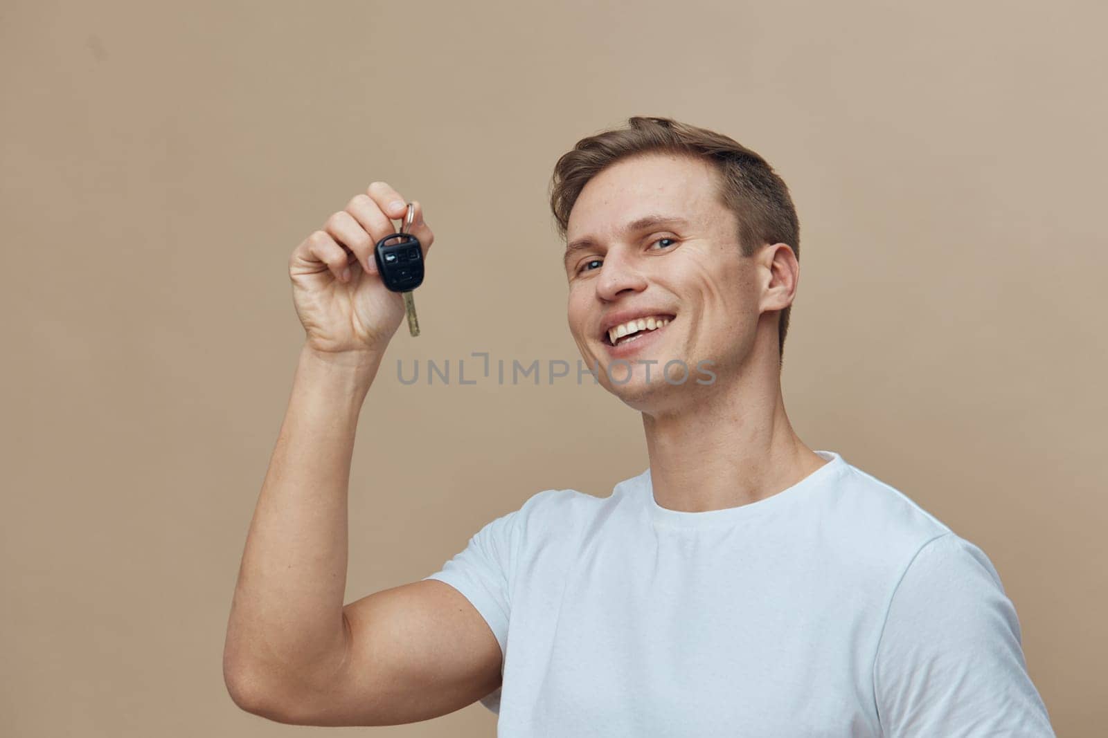 Cheerful young man in a white t-shirt smiling and holding car key, standing against a plain beige background, representing happiness, success, and achievement. Lifestyle and automotive concept