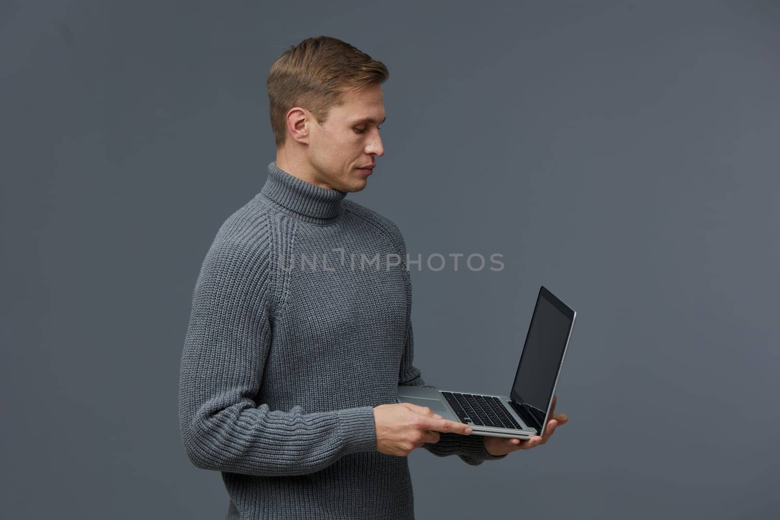 young caucasian man working with laptop using AI chat technology wearing cozy gray sweater isolated on plain gray background showing focus on digital communication and artificial intelligence tools concept