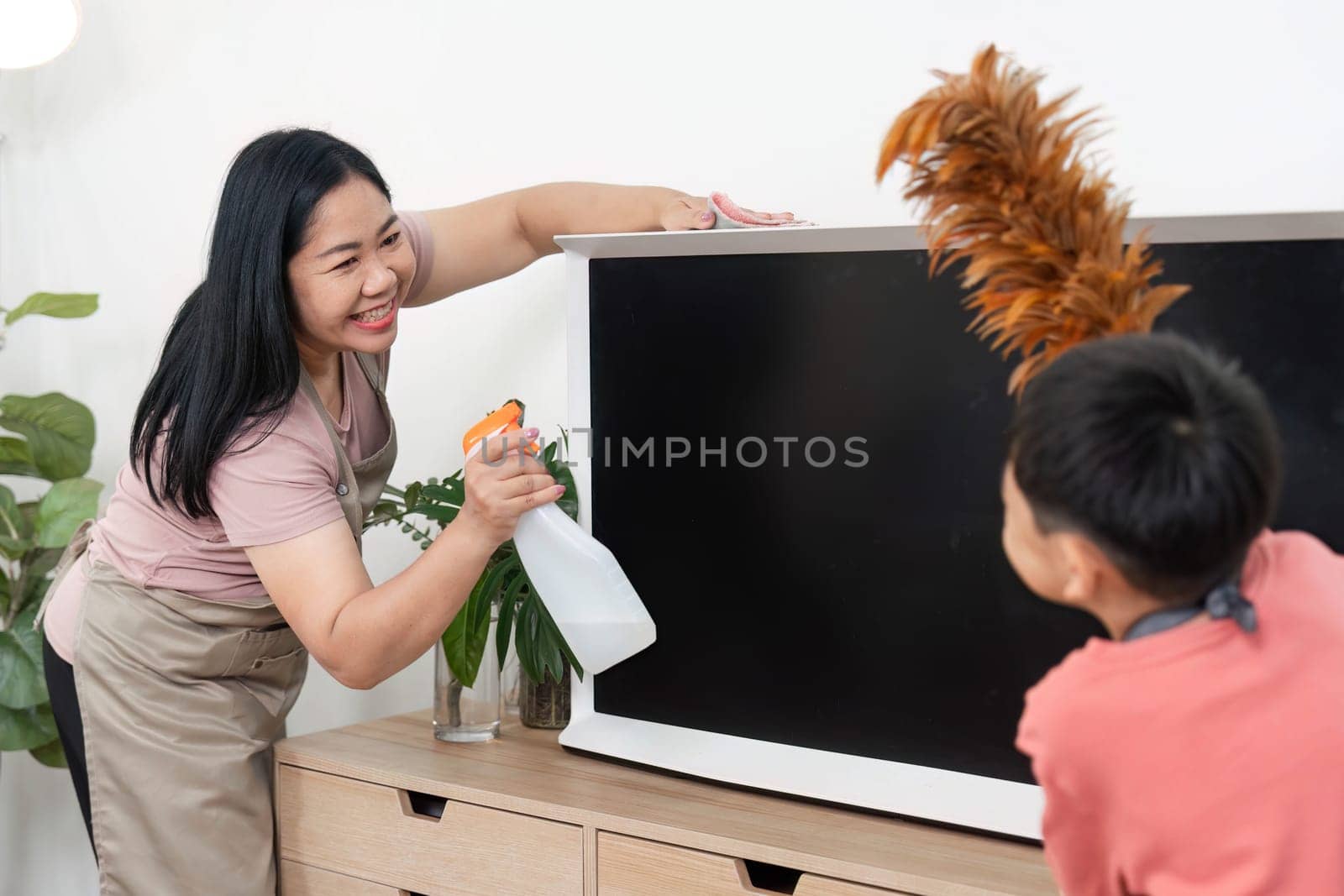 A joyful interaction between a woman and a child as they clean together, using a spray bottle and feather duster to make home cleaning a fun activity.