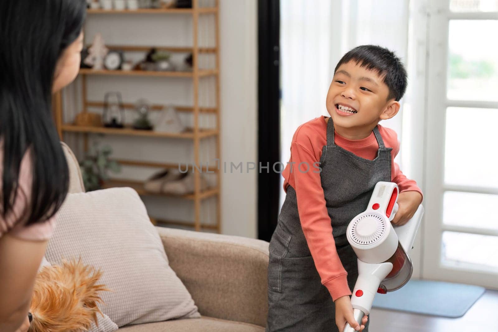 A young boy joyfully assists his mother with cleaning the living room, showcasing teamwork and responsibility in a family setting. The image captures a moment of fun and learning in household chores.