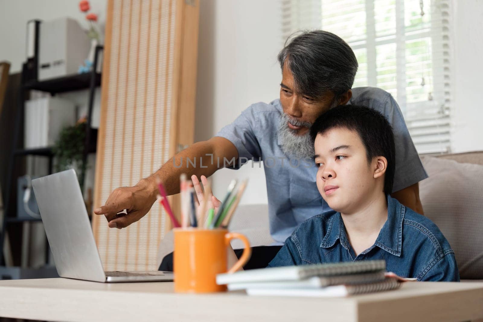 A grandfather supports his grandchild with Down syndrome while using a laptop for learning, highlighting the importance of technology in education.