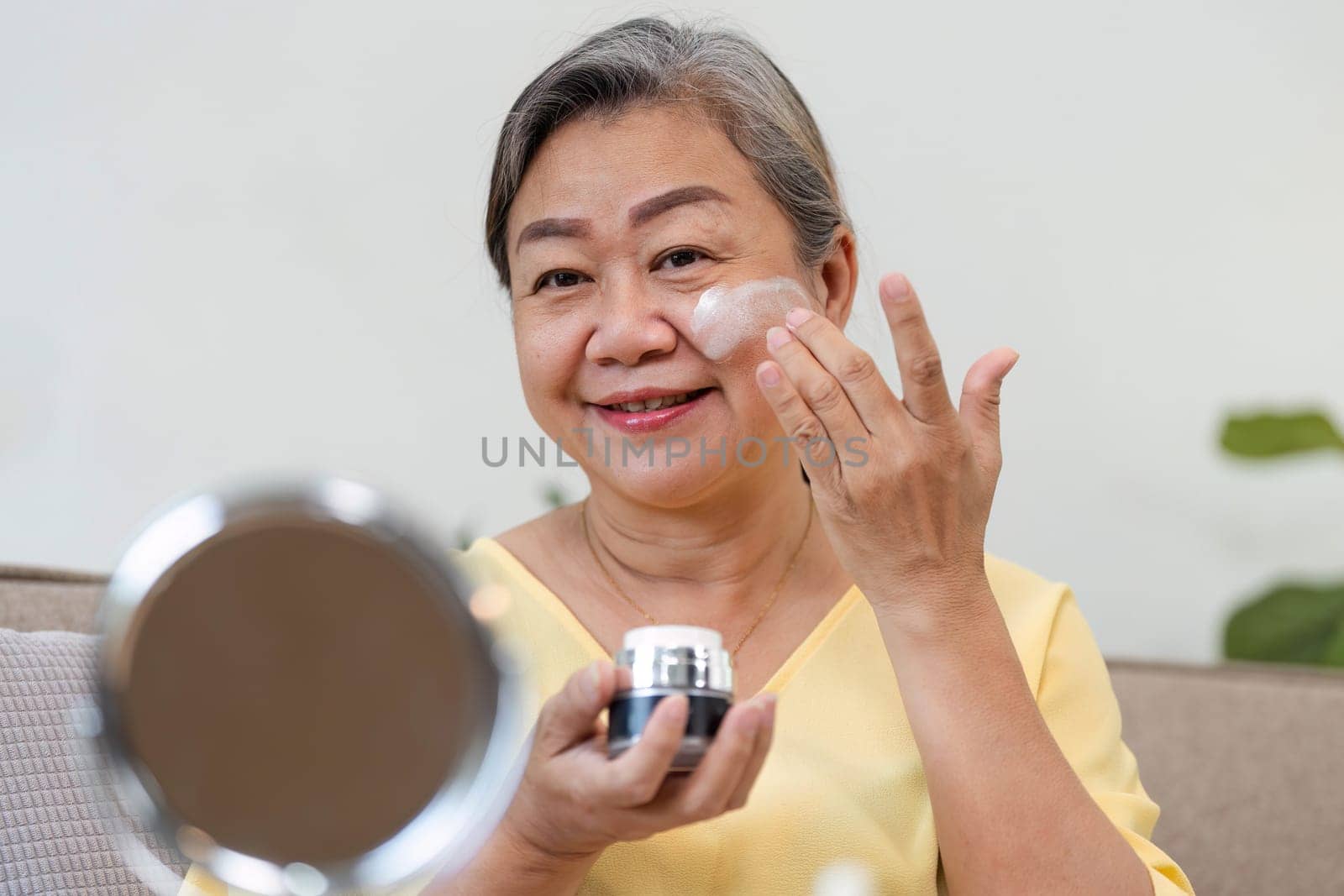 An elderly woman smiles as she applies skincare cream, showcasing the joy of self-care and the beauty of aging gracefully in her cozy home.