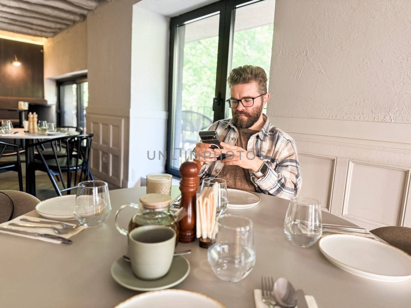 Handsome man browses his smartphone while waiting for coffee. Digital routine, casual lifestyle and urban cafe culture.