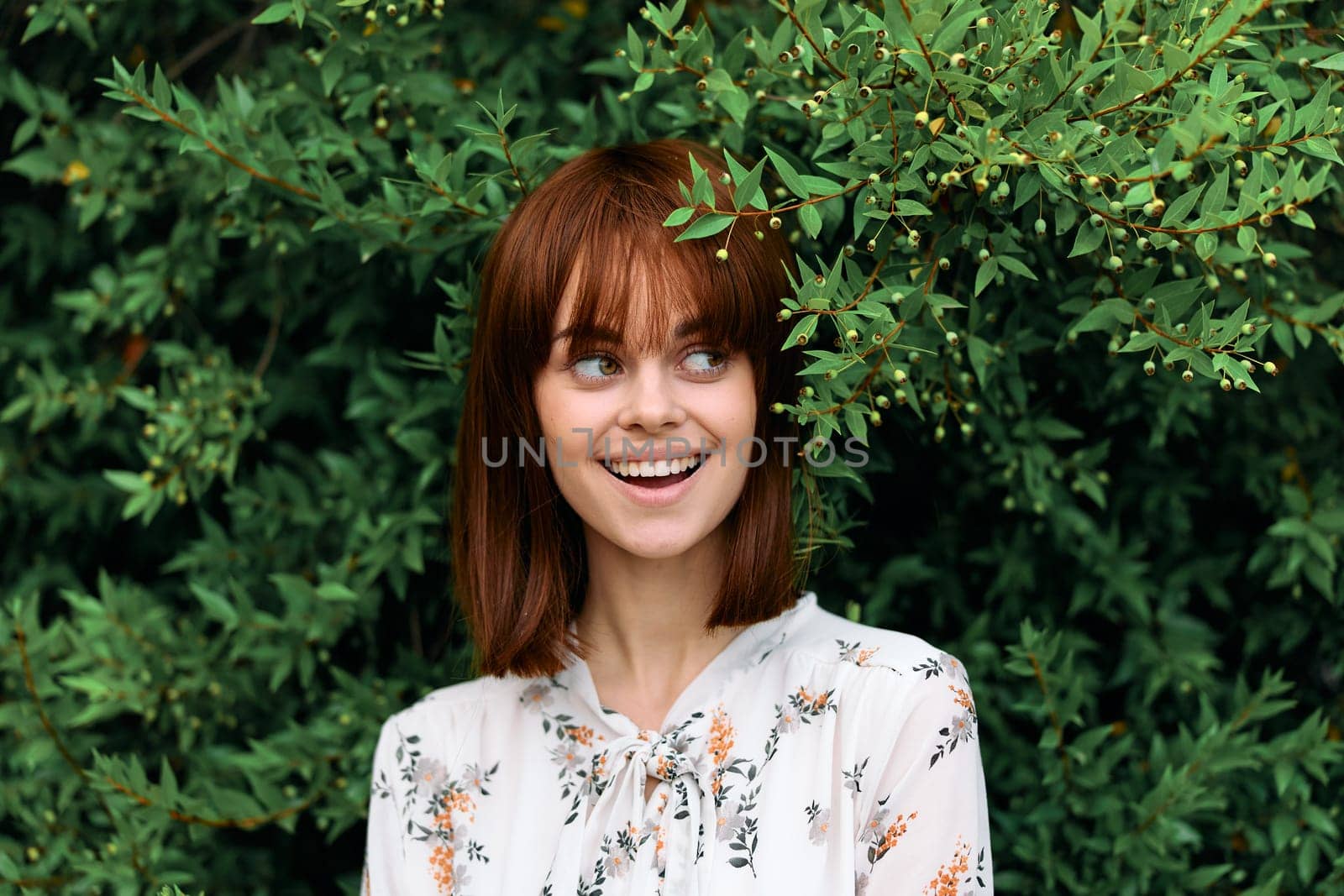 Vibrant woman with red hair posing in front of lush green plants and bush