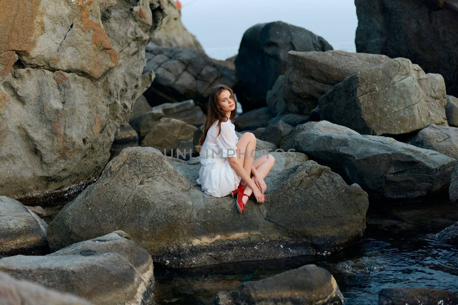 Serene woman in white dress and red shoes sitting on rocky outcrop in the middle of the ocean