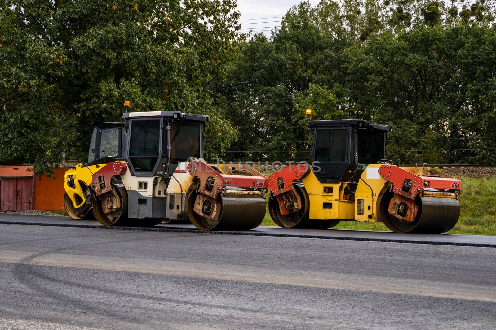 Two heavy-duty road rollers positioned side by side on a freshly laid highway. The scene highlights machinery used in asphalt compaction and infrastructure development amid a green backdrop.