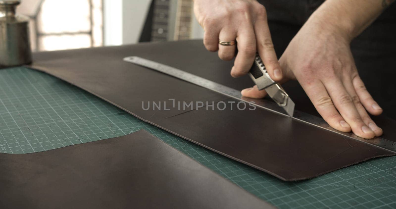 A close-up of a leatherworker using a utility knife and metal ruler to cut straight lines into a thick sheet of brown leather. The scene showcases detailed craftsmanship and tool precision on a gridded cutting mat.