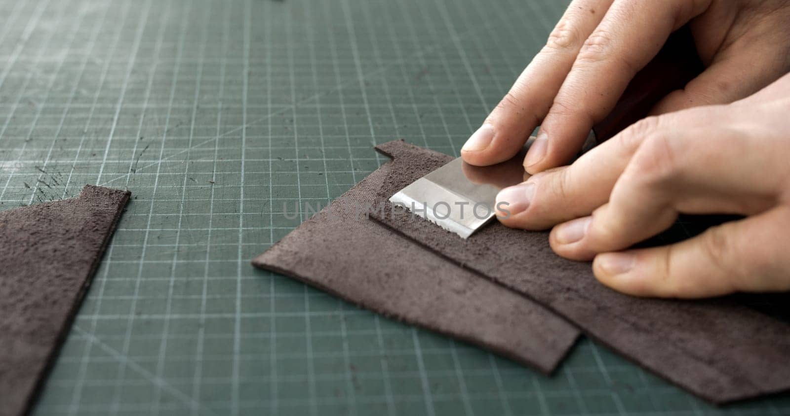 Close-up of a leatherworker's hands using a metal skiving tool to thin the edge of brown leather pieces on a cutting mat. The image captures the precision and delicacy required in crafting high-quality leather goods.