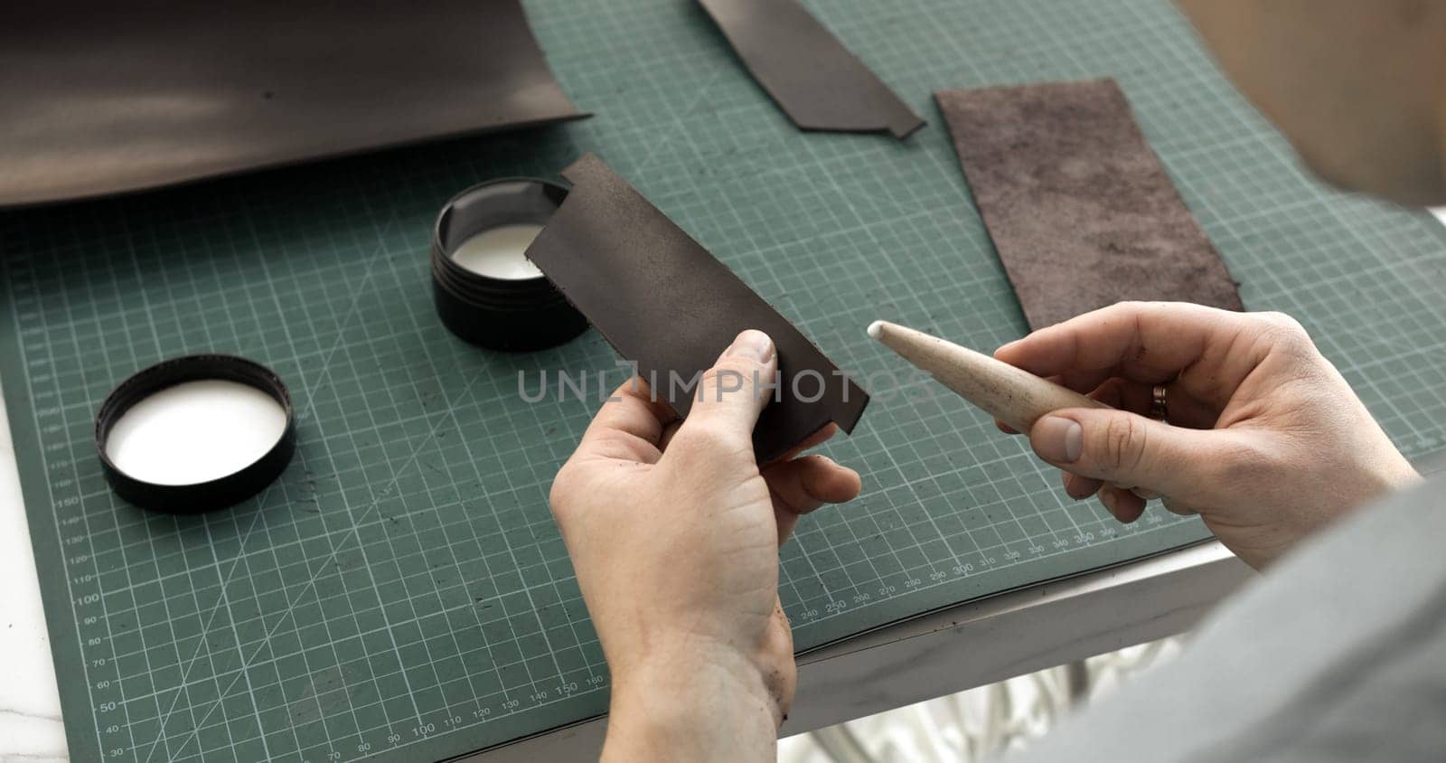 A leatherworker holds a strip of leather in one hand and a bone folder in the other, preparing the edge for burnishing. Surrounding tools and leather pieces are visible on the green gridded work surface.