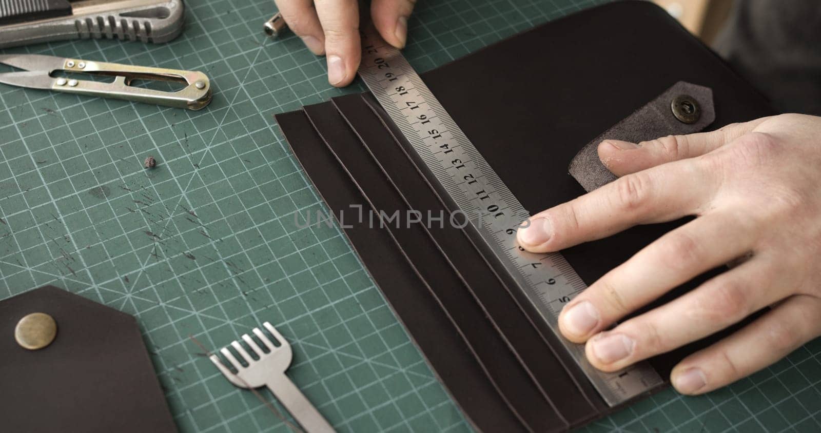 Close-up of a craftsman measuring leather pieces with a metal ruler on a green cutting mat. Tools and crafting materials surround the workspace during precise leathercraft work.