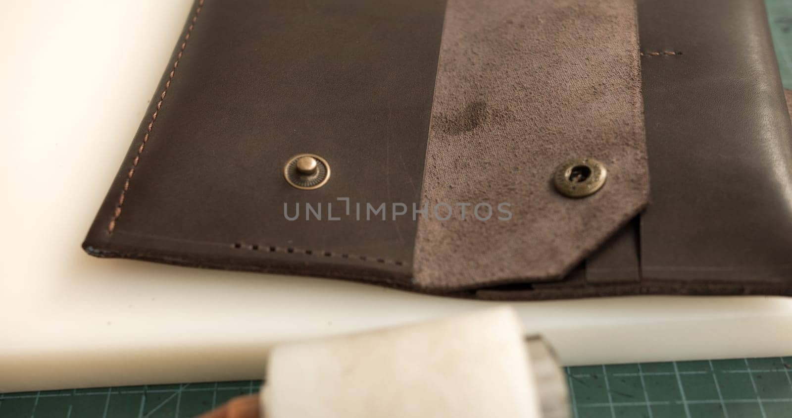Detailed close-up of a handmade leather wallet with visible stitching and a snap button. The wallet is placed on a smooth white surface, showing craftsmanship and clean finish.