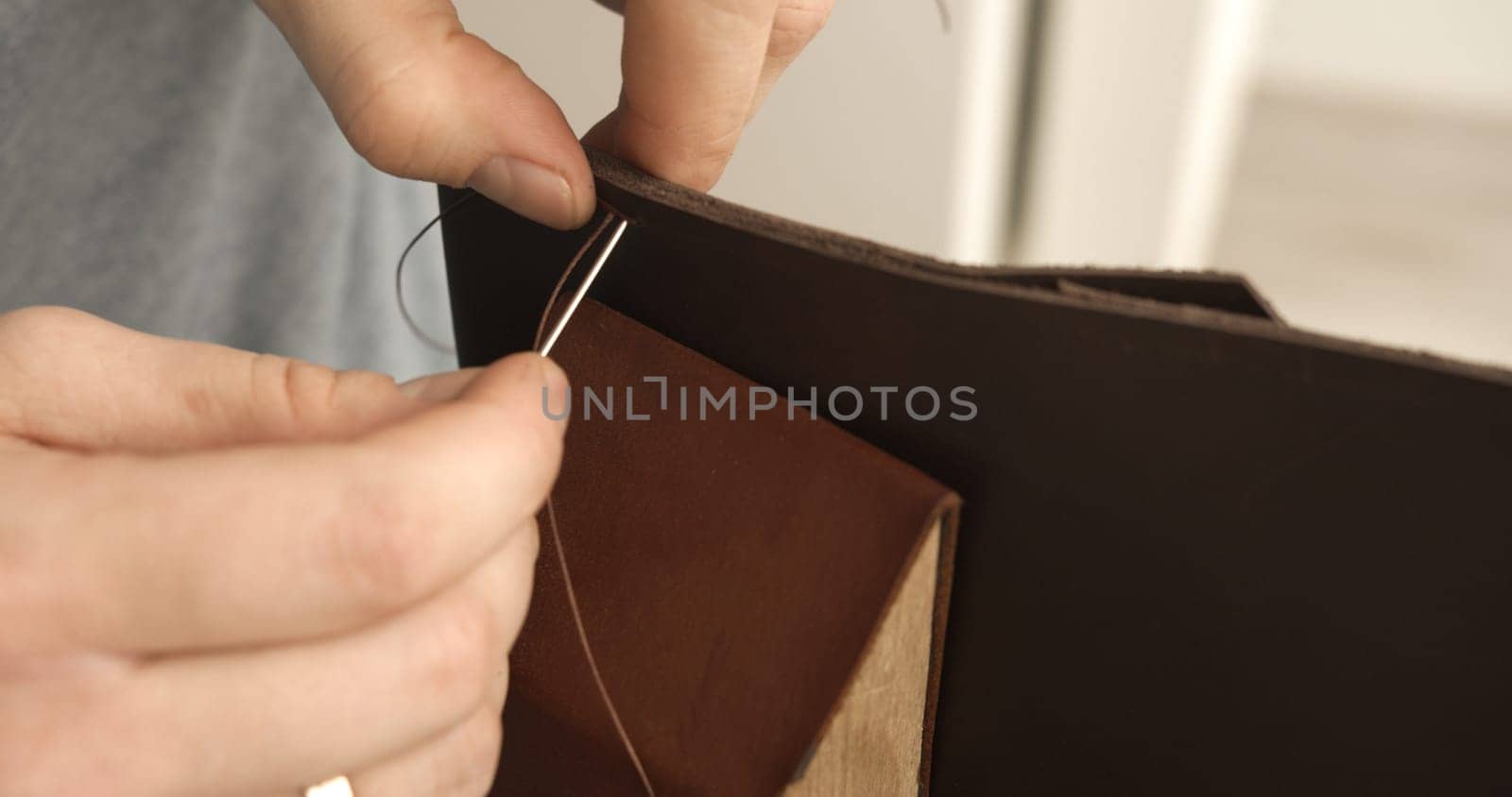 Close-up of leatherworker's hands stitching a brown leather flap using a needle and thread. The detailed view showcases precision in handmade leather crafting during wallet production.