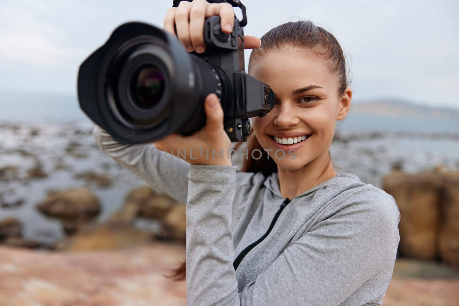Happy young woman in a gray sweatshirt capturing moments with her camera at the beach, radiating enthusiasm for photography and outdoor experiences
