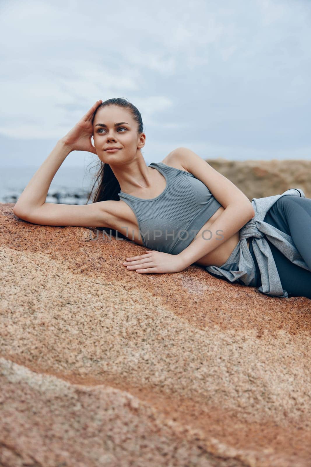 A fit young woman in gray sportswear rests on a rock near the sea, enjoying the calm atmosphere and showcasing a lifestyle of health and wellness
