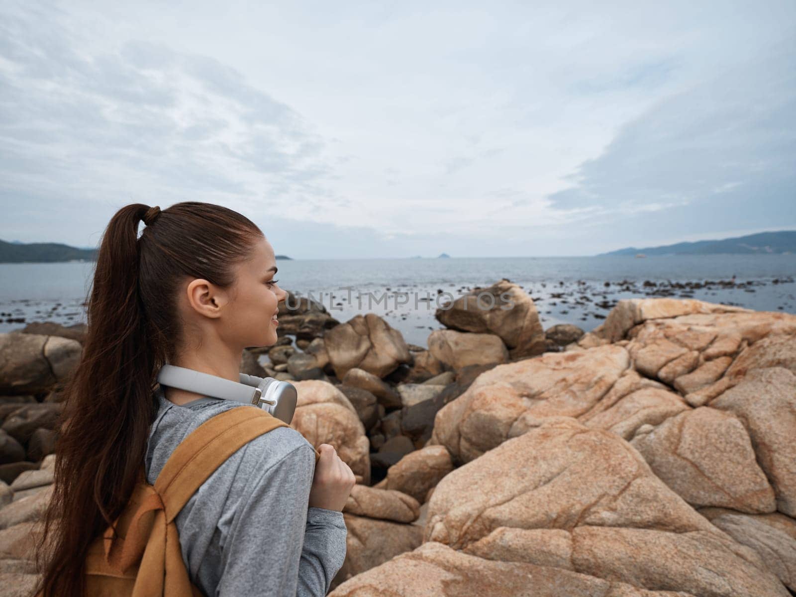 A thoughtful young Asian woman dressed in casual attire stands on rocky terrain, looking towards the ocean, embodying adventure and serenity in a picturesque coastal setting