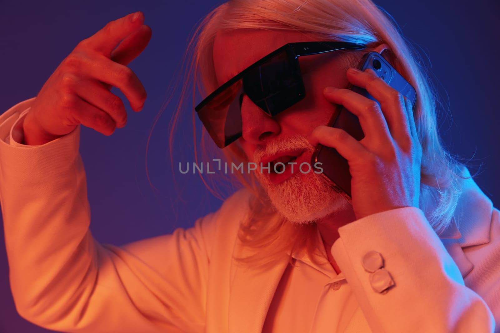 A fashionable older man with long white hair and sunglasses is engaged in a lively phone conversation The background features striking blue and purple hues, adding to the trendy and energetic vibe of the image