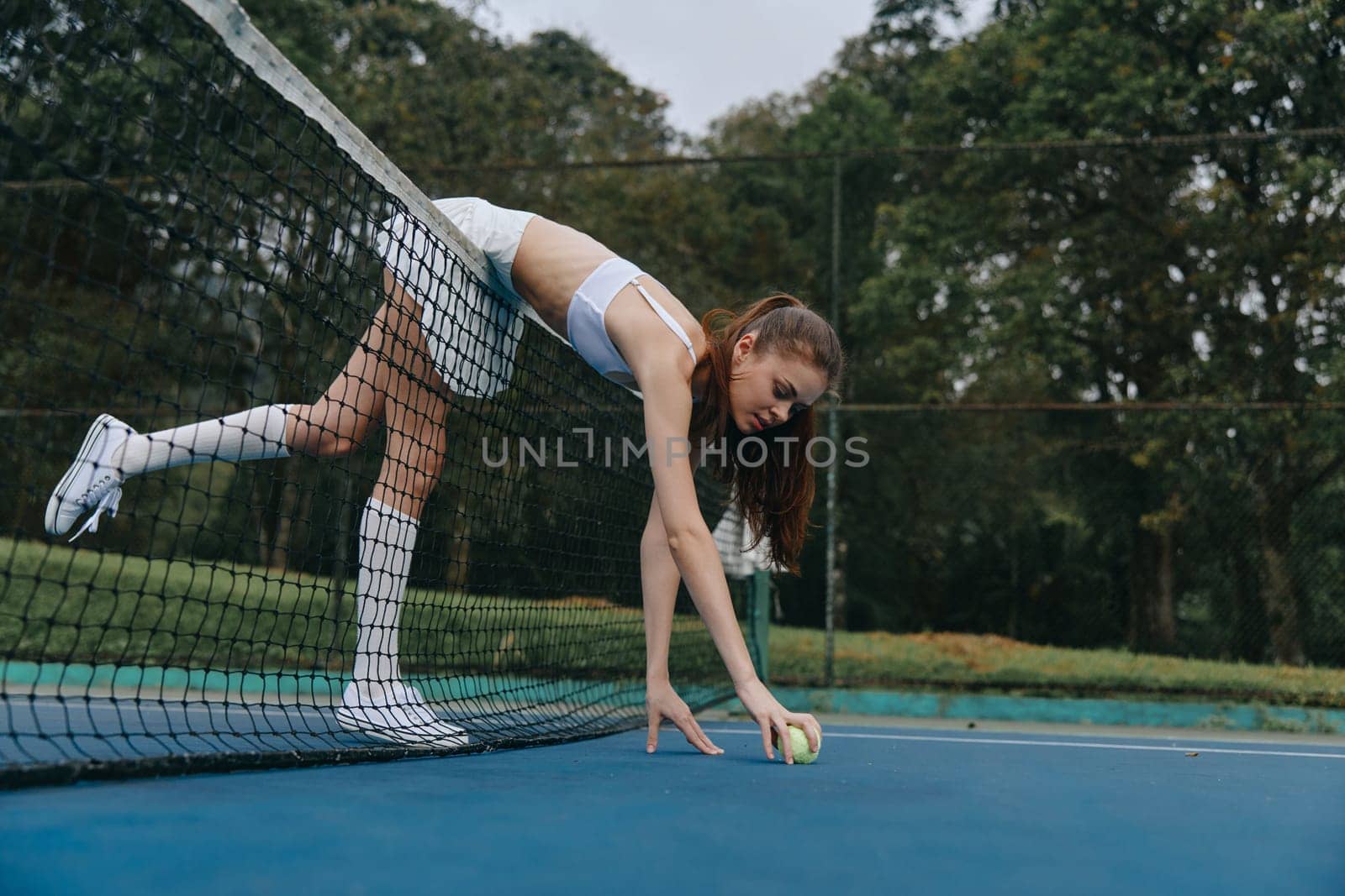 Determined young tennis player in white sportswear bending to pick up a ball on a blue court, surrounded by lush greenery, reflecting athleticism and summer sports spirit
