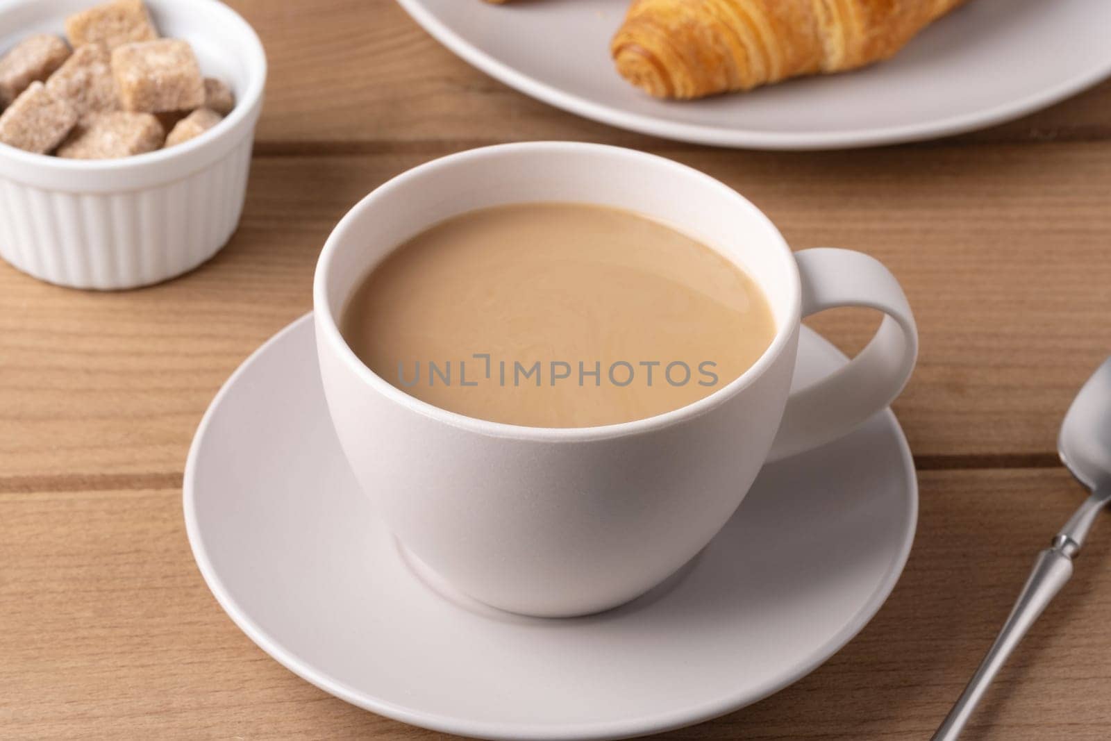 A white cup filled with warm coffee is placed on a saucer next to a croissant and a small bowl of sugar cubes on a wooden table. The setting suggests a cozy breakfast.
