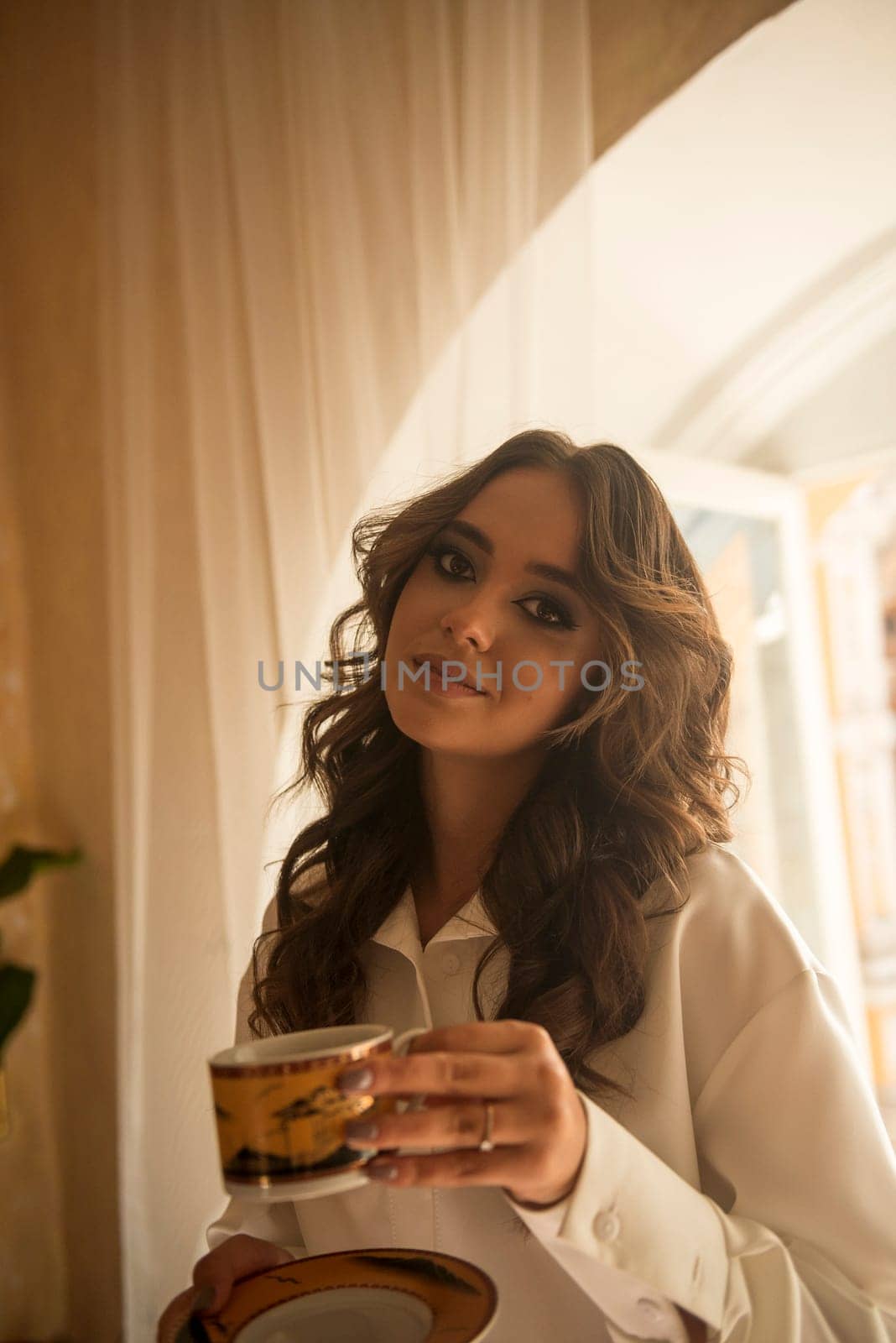 young romantic brunette with long wavy hair in a room with a cup and saucer in her hands. dressed in a white silk suit. looks at the camera with a slight smile. close-up