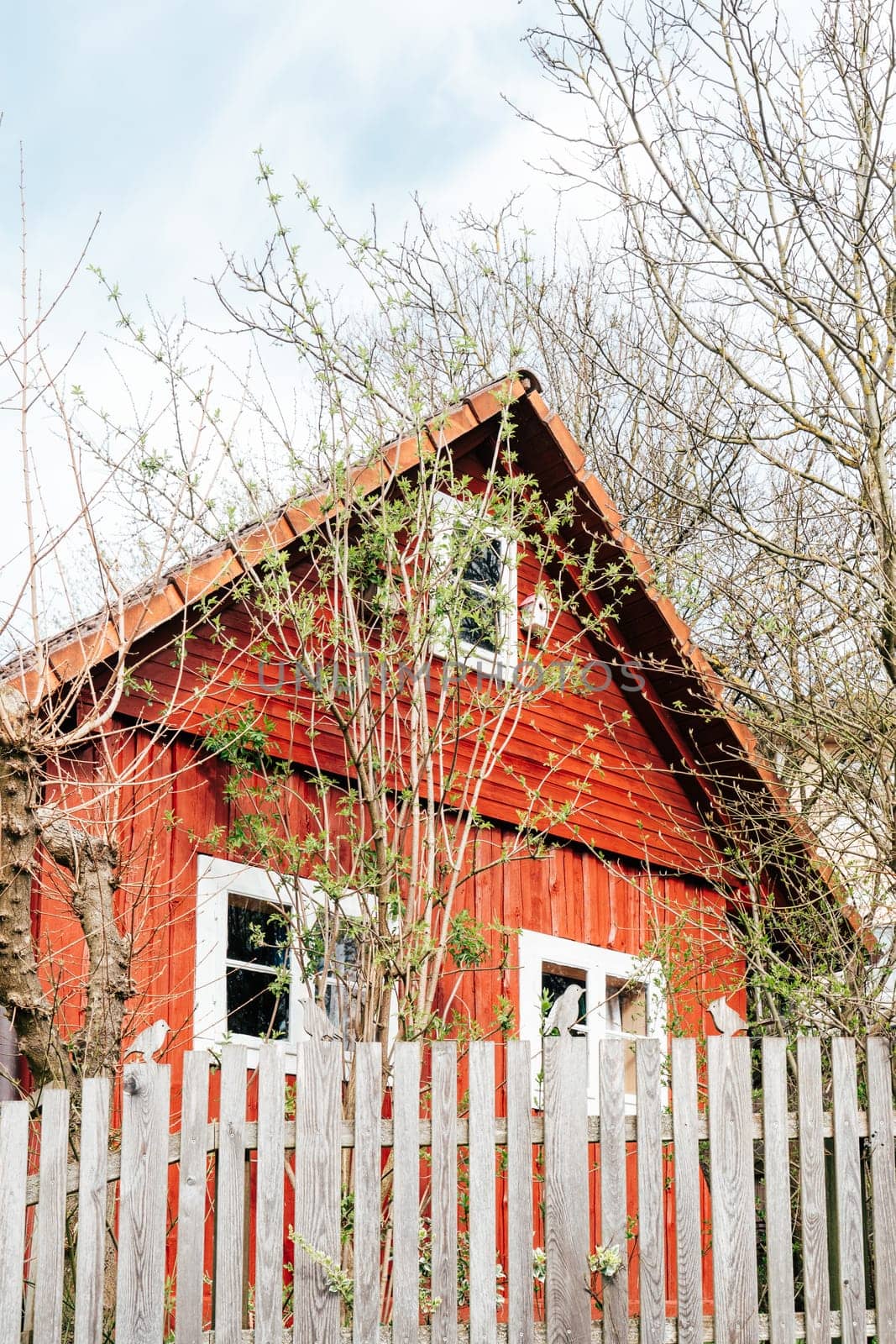 A quaint red house with a triangular roof sits amidst budding trees and shrubs. Wooden fences encircle the property, highlighting the peaceful charm of early spring in a rural setting.