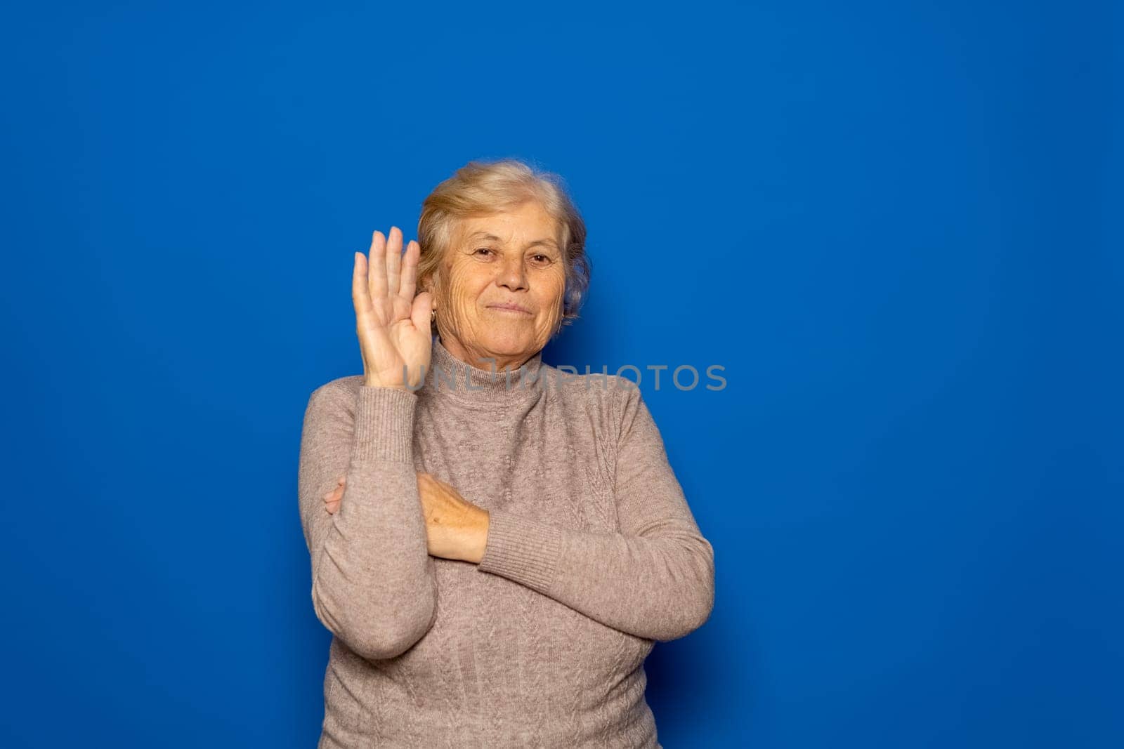 Hello concept. A happy, smiling, elderly Caucasian woman in her 70s waves with her hand outstretched. Isolated on a blue studio background