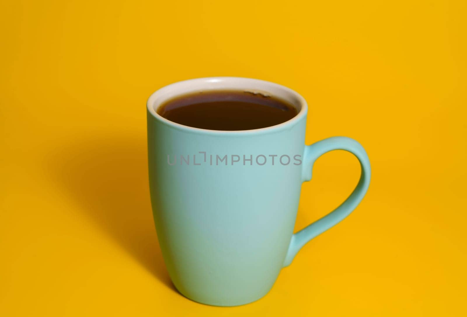 A matte blue ceramic mug filled with black coffee placed on a solid yellow background in a minimal studio setting.