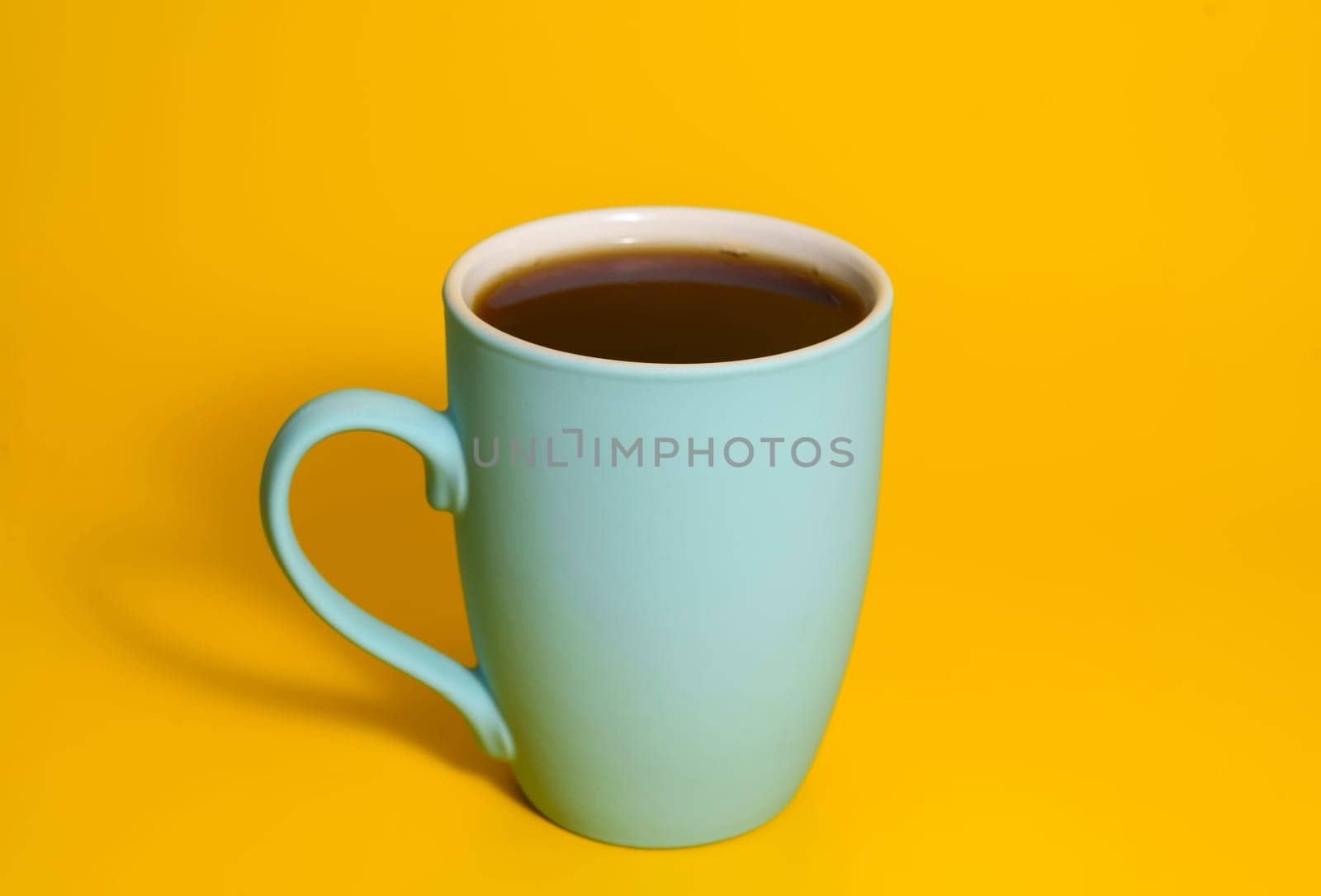 A turquoise ceramic mug filled with black coffee placed on a solid bright yellow background in a minimalist studio setup.