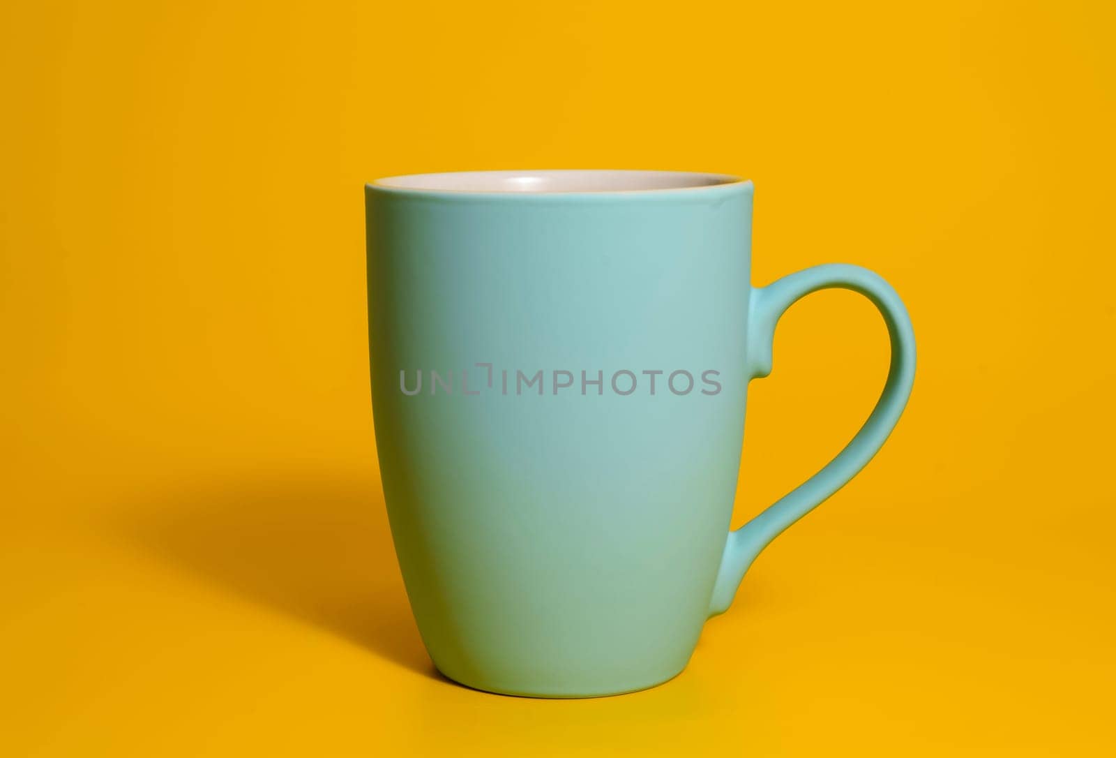 A single matte blue ceramic mug with a handle is placed against a solid yellow backdrop, minimal and clean studio lighting.