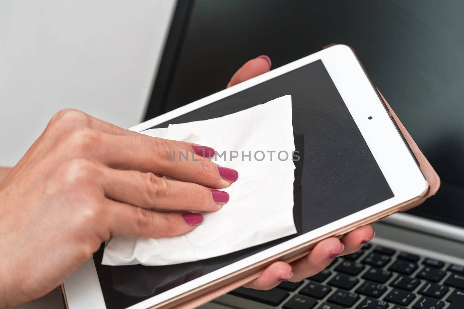 Woman cleaning tablet device with cotton paper tissue, wiping black screen, closeup detail. Blurred laptop keyboard background