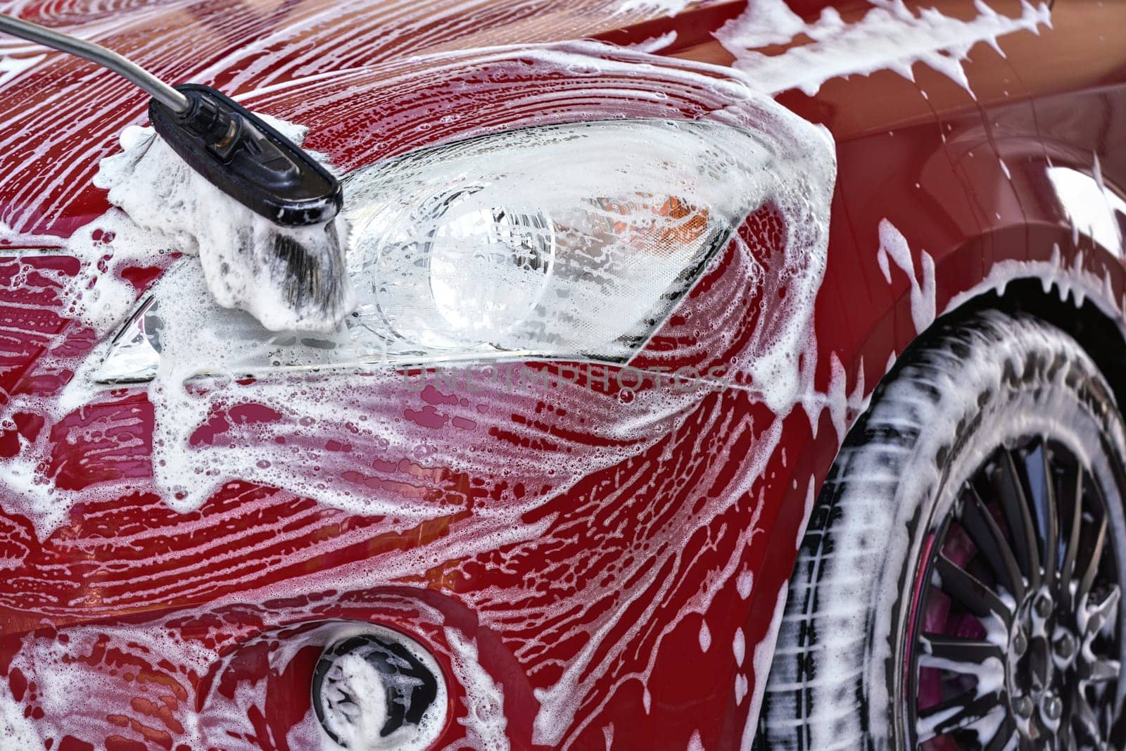 Front head light of red car washed in self serve carwash, brush leaving white soap strokes on glass, closeup detail