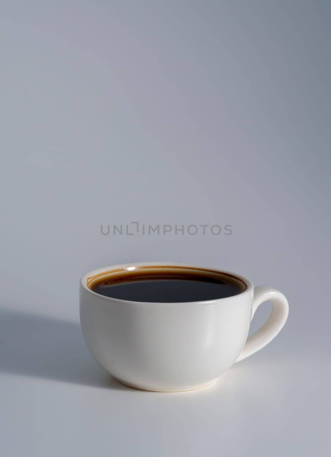 A minimalist white ceramic cup filled with black coffee, placed on a soft gray surface with gentle lighting and subtle shadow.