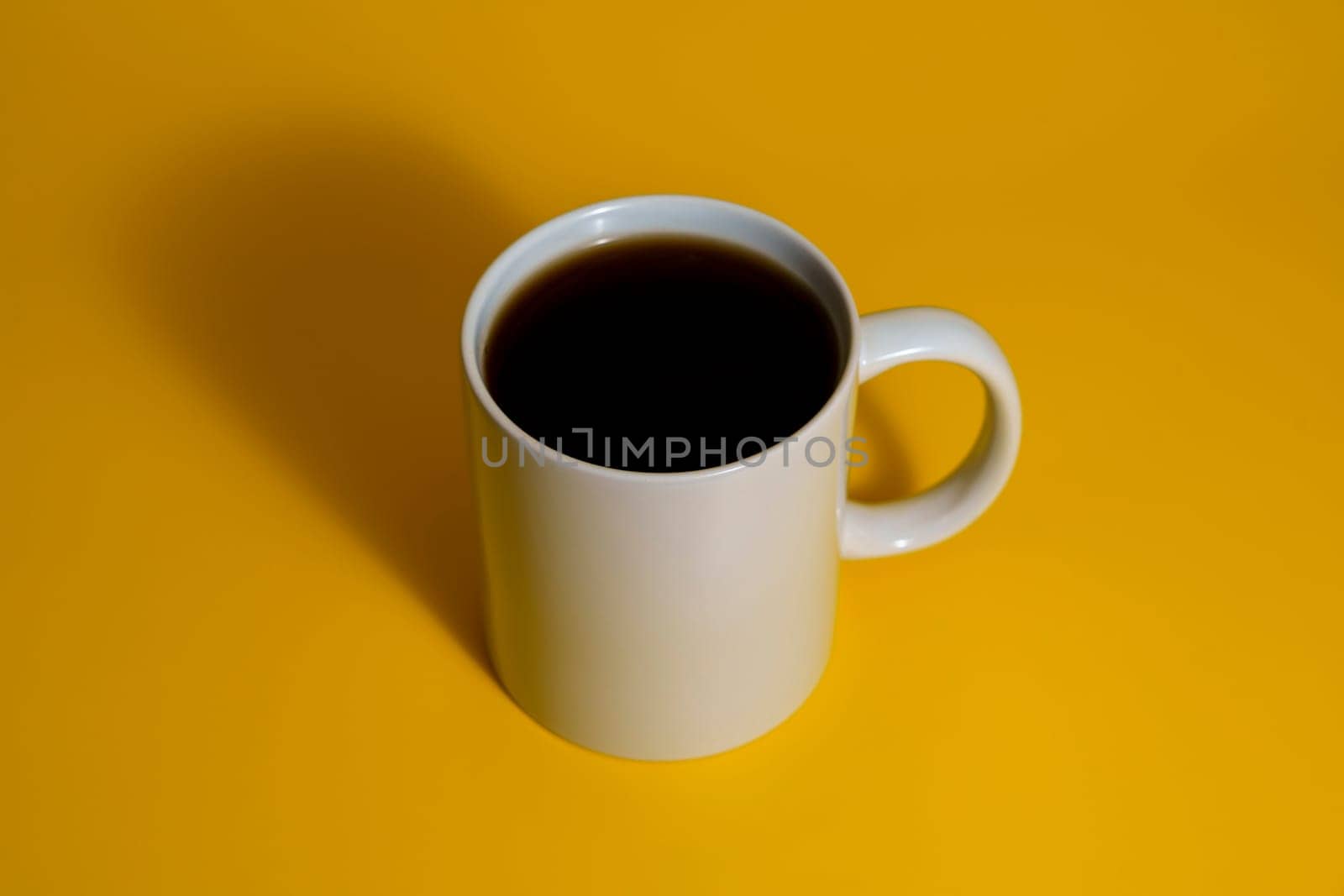 Top side view of a white ceramic mug filled with dark coffee placed on a vibrant yellow surface with dramatic shadow.