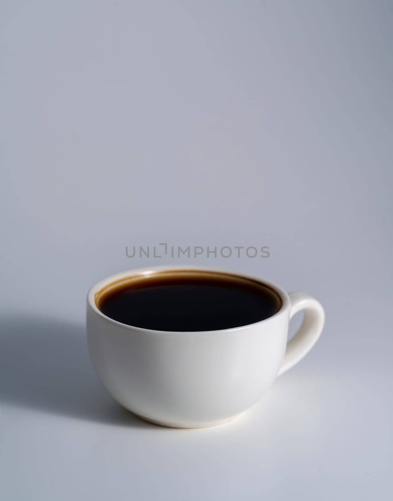 Minimalist white ceramic cup filled with black coffee, placed on a clean light gray surface with soft natural lighting and gentle shadows.