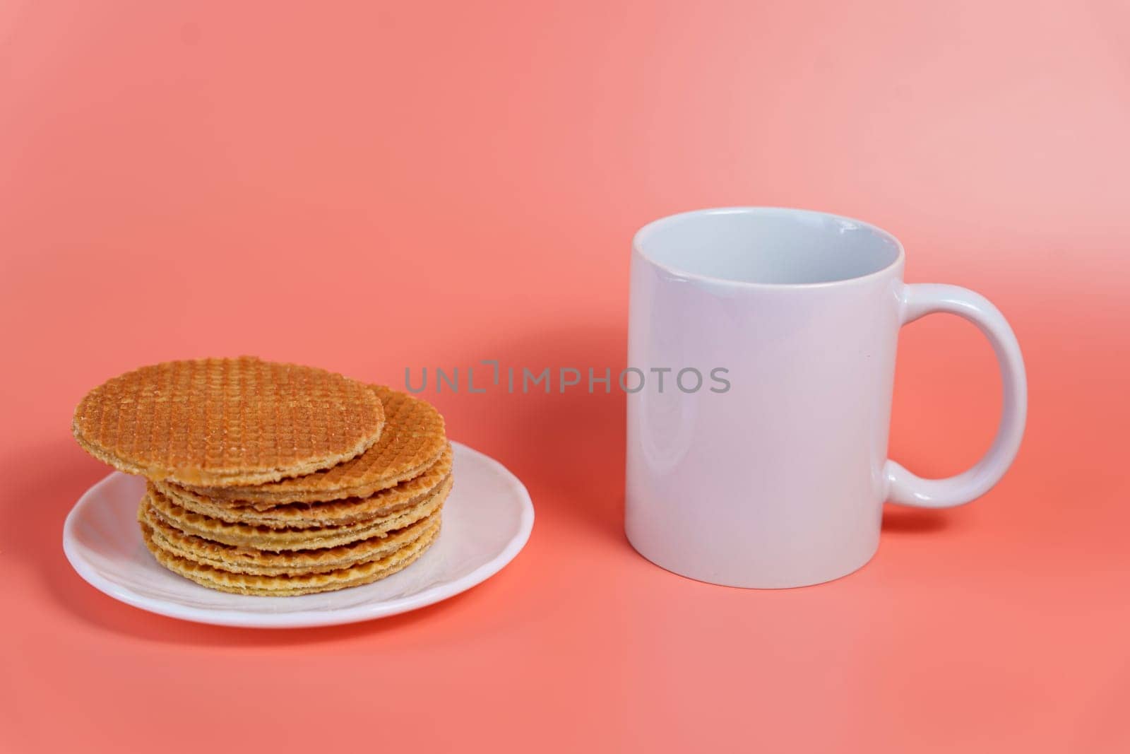 Traditional Dutch stroopwafels stacked on a white plate next to an empty white mug over a pastel pink background.