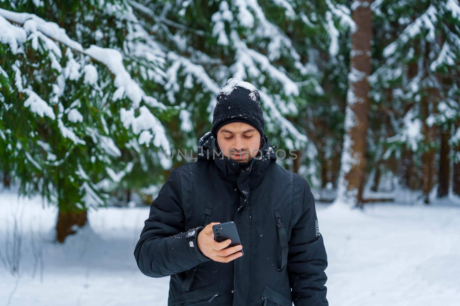 Young man in black winter coat and beanie standing in a snow-covered forest, looking at smartphone with snow on his hat.