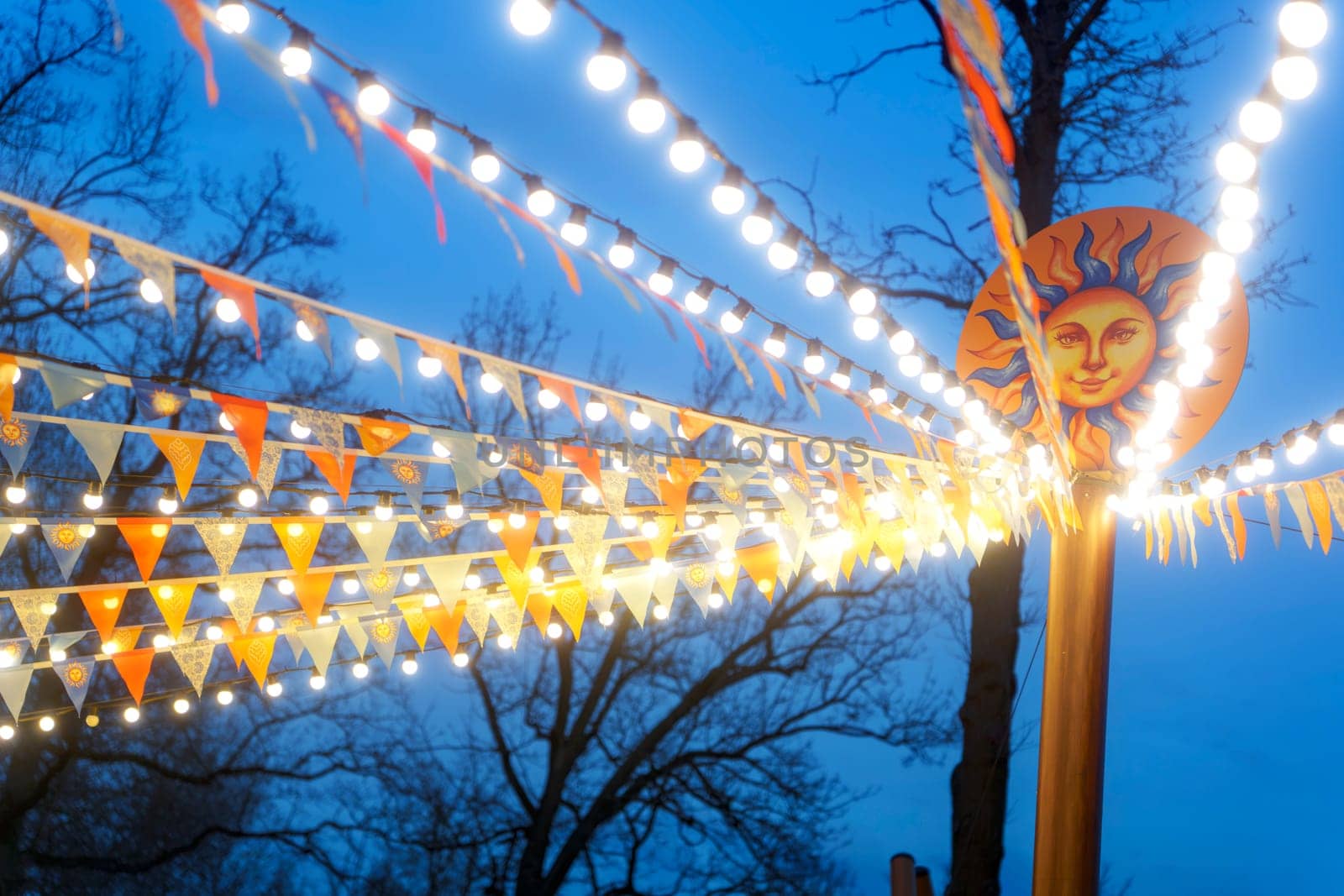 Bright string lights and triangular flags stretch from a sun face pole at an outdoor festival during twilight, set against a deep blue sky and bare trees.