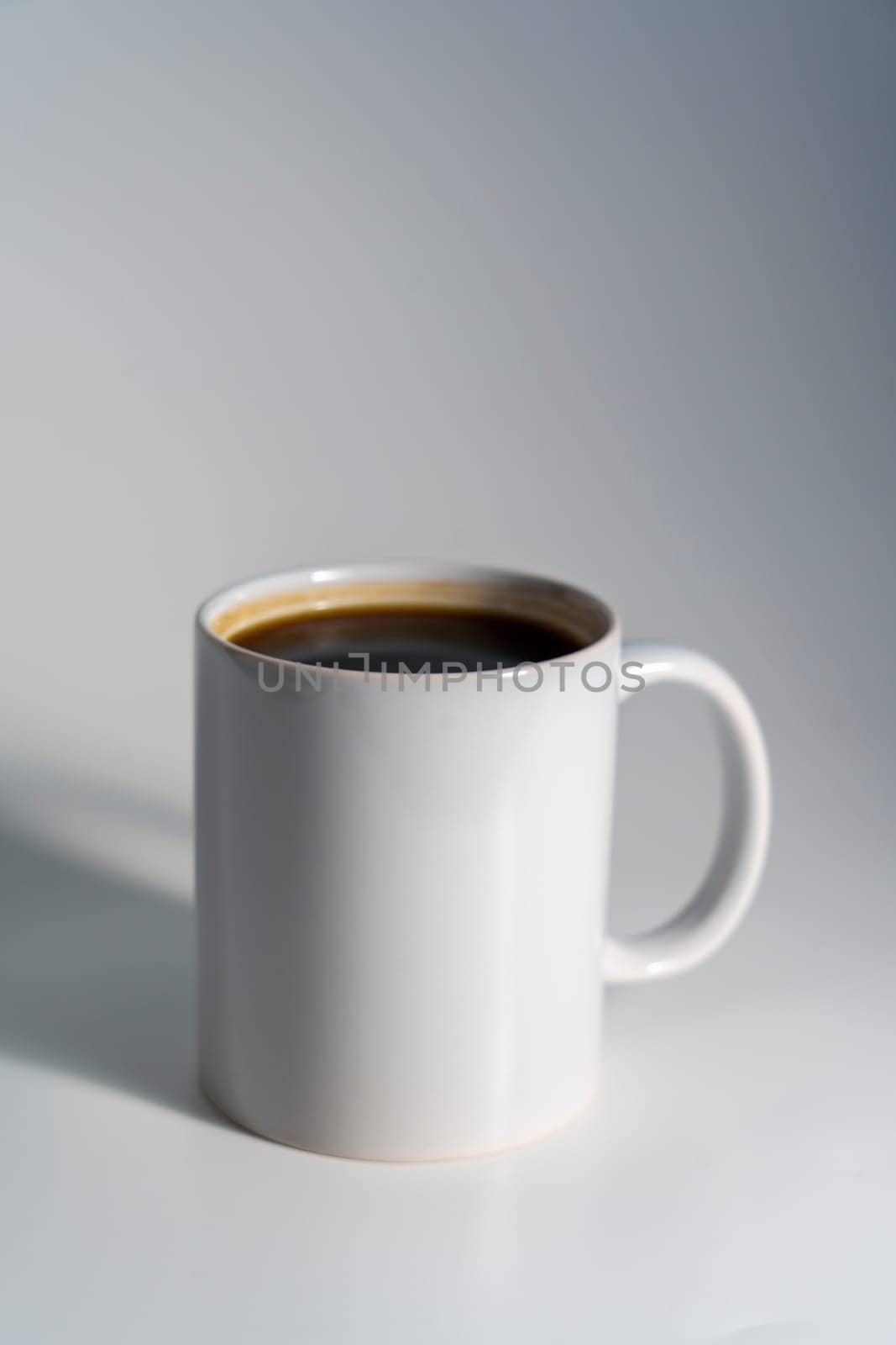 A simple white ceramic mug filled with coffee, placed on a clean light background with soft natural lighting and gentle shadow.