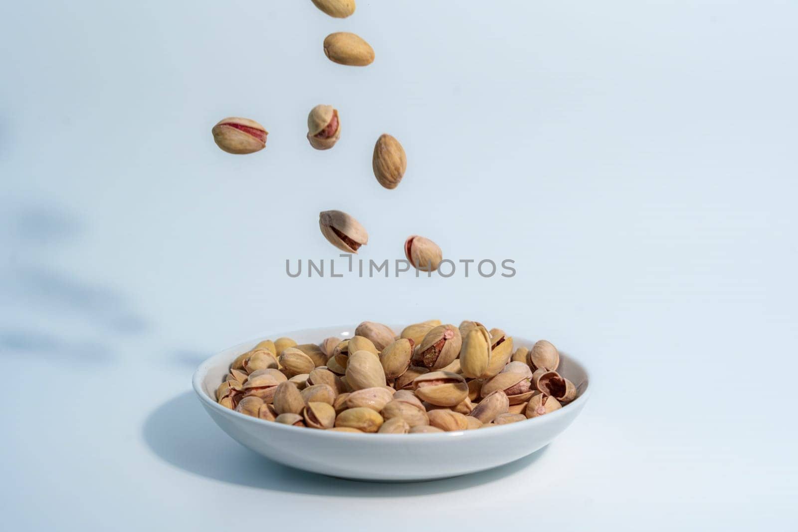 Pistachio nuts falling into a white ceramic bowl already filled with pistachios, captured against a clean light blue background.